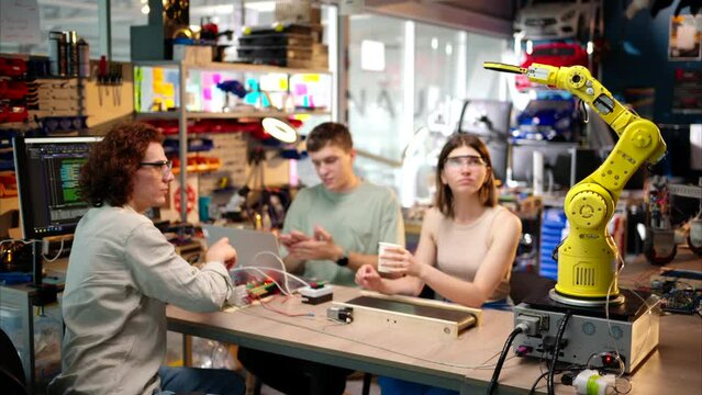 Young happy engineers programming an yellow robotic arm in the workshop to grab cardboard water glass, computer programming training for coffee preparation, celebrating success