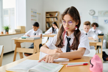 Schoolgirl sitting at her desk and reading textbook during lesson in school