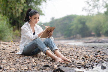 Photo of young attractive Asian woman writing on diary while sitting outdoors in the park, Writer...