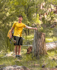 a young man in a yellow T-shirt leaning against the trunk of a fallen tree with a guitar in his hand