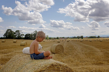 Beautiful blond child, boy, lying on a haystack in the field. Amazing landscape, rural scene with clouds, tree and empty road summertime, fields of haystack next to the road