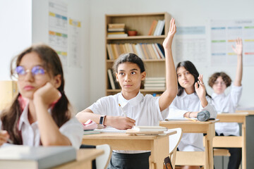 Schoolgirl sitting at her desk and raising her arm, she answering at lesson at school
