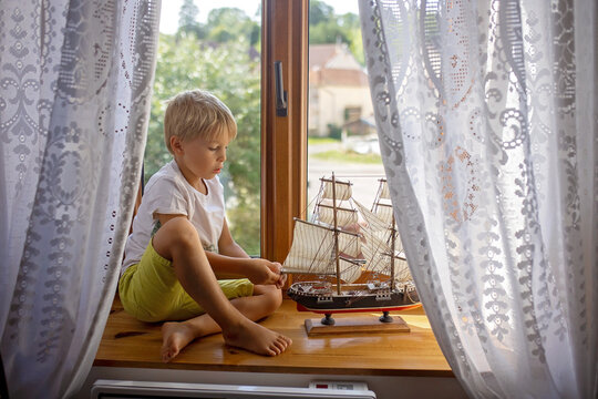 Cute blond child, boy, playing at home with big ship on the window