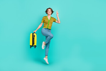 Fototapeta na wymiar Full length portrait of excited person jumping hold suitcase show v-sign isolated on turquoise color background