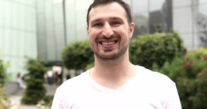 Positive guy smiling at camera. Portrait of male model posing on camera in green park. Cheerful man face with positive emotion outdoors.