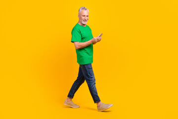 Full size profile photo of cheerful aged man walking hold smart phone typing blog post isolated on yellow color background
