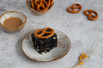 	
Dark chocolate brownies with salted caramel and pretzels on a grey stone background. Top view. Dessert from American cuisine.
