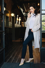 Stylish young female with dark hair, white coat, black trousers, and heels, posing against cafe background, chatting over the phone with hand in pocket.
