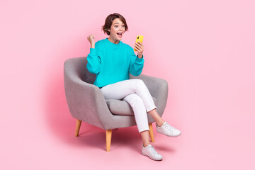 Full size photo of pretty young girl sit armchair raise fist hold telephone dressed stylish blue...