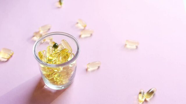 Transparent yellow capsules with vitamins are poured into a transparent glass standing on a pink background. Fish oil supplement. The concept of health, vitamins, and medicine. Omega 3, vitamin D