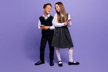 Photo of positive schoolkids blogging about school courses on smartphones isolated purple color background