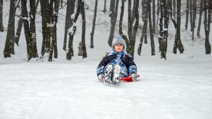 Fototapeta na wymiar Boy gleefully gliding down a snow-covered hill on his plastic sled, grinning from ear to ear. The perfect image of winter holidays, fun in the snow, and the joy of childhood.