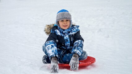Fototapeta na wymiar Portrait of happy cheerful boy sitting on plastic sleds and smiling in camera during snowfall