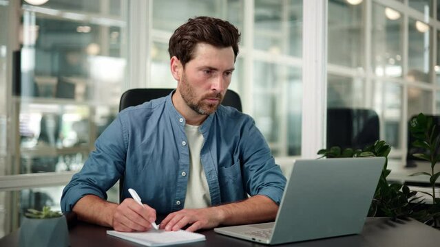 Serious businessman sitting in front of laptop at office desk, taking notes in business documents working on startup or project. Manager preparing for presentation, checks documents, does paper work.