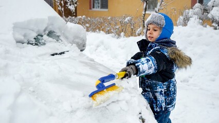 Young boy happily clearing snow off his parent's car with a brush on a cold winter morning. Concept of responsibility and vehicle maintenance. - obrazy, fototapety, plakaty