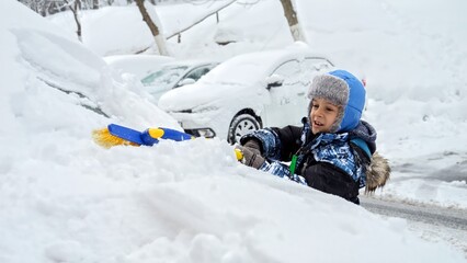 Cheerful boy cleaning snow from a car with a brush after a winter storm. Concept of helping out...