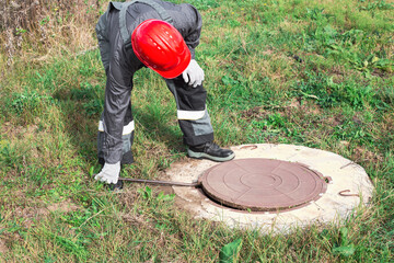 A worker in overalls and a helmet opens a manhole with a crowbar. Control and maintenance of sewerage, septic tanks and plumbing
