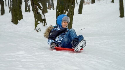 Happy smiling boy riding down the snowy hill on plastic sleds. Concept of winter holiday, children having fun and playing outdoors in snow, Christmas vacation.