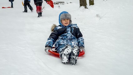 Fototapeta na wymiar Cheerful young boy sledging down a snowy hill on his plastic sleds, exuding pure joy and enthusiasm. Holiday season and the fun children