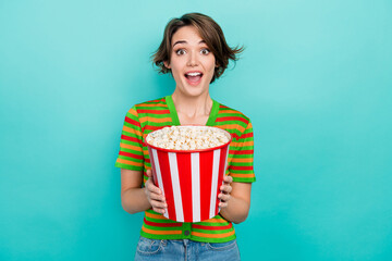 Portrait of astonished cheerful girl hands hold big popcorn bucket isolated on turquoise color...