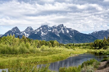 a grassy field with trees in the background and mountains towering over the river - Powered by Adobe