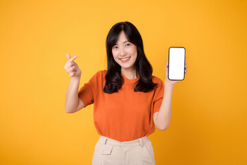 Cheerful young Asian woman in her 30s, wearing an orange shirt, showing mini heart hand gesture and smartphone blank display screen on yellow background. new application technology concept.