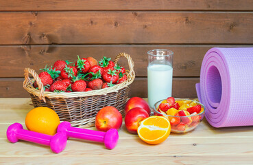 Fresh strawberry, yogurt, healthy fruit salad, fresh fruits, dumbbells and yoga mat on wooden surface.