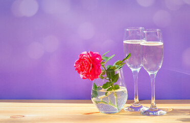 Pink rose and  two glasses of champagne on purple background. Greeting card with space for your text.