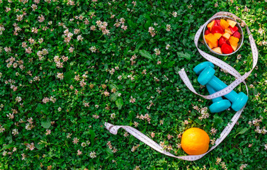 Dumbbells, measuring tape and fruit salad on  green grass.  Top view, copy space.