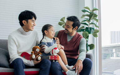 Asian male Gay couple taking care adopted little daughter girl, playing, smiling with happiness, sitting on sofa in living room at home. LGBT, Family and Kid concept.