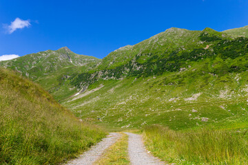 Empty dirt and gravel road with grass covered mountains in the background and clear blue sky