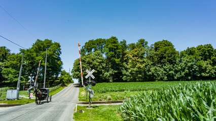 View of An Amish Horse and Carriage Traveling Down a Rural Country Road After Crossing a Rail Road Crossing on a Sunny Spring Day