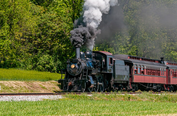 Fototapeta na wymiar A View of an Antique Restored Steam Passenger Train, Approaching, Blowing Black Smoke, Traveling Thru Rural Countryside on a Sunny Day