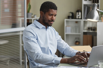 Portrait of handsome black businessman working with laptop at desk in office, copy space