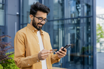 Young successful satisfied hispanic man smiling outside office building, businessman using app on phone for online shopping in online store, man holding bank credit card and smartphone. - obrazy, fototapety, plakaty