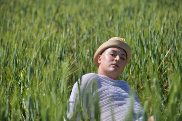 Non-binary person, young and South American, very makeup, wearing a hat, posing sitting in the middle of a field of green wheat. Concept queen, lgbtq+, pride, queer.