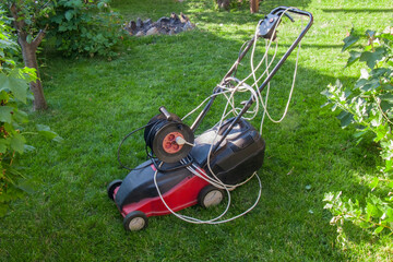 Fototapeta na wymiar red electric lawn mower on a green lawn with wires and an electric extension cord, portable socket