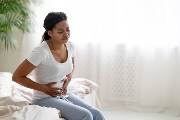 Black woman suffering stomach pain, sitting on bed and touching belly