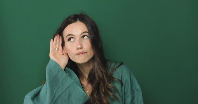 Cute curious woman keeps hand near ear tries to overhear something very interesting listens somebody secret looks camera likes collecting rumors poses against green background. Eavesdropping concept.