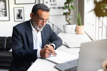 Obraz premium Busy serious caucasian senior businessman in suit makes notes in contract and looks at computer