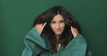 Brunette curly woman with hair under ears looks shy next fix hair and start smiling, look proud standing isolated on green background. from shy to confident.