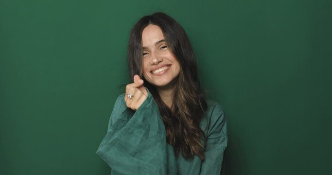 Smile curly brunette woman making finger heart sign, Korean small heart finger sign. Love concept. Romantic lady making heart sign with fingers and smiling isolated on green background.