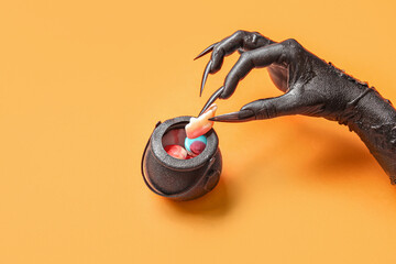 Black hand of witch with cauldron and tasty candies on orange background, closeup. Halloween...