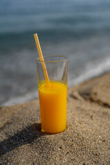 a glass of orange juice with a straw stands on the sand against the backdrop of the sea