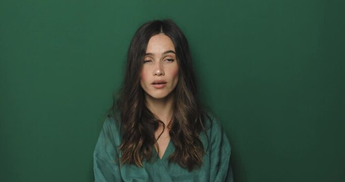 Displeased young woman show shut up gesture standing on green background and looking at camera. Angry woman don't want listen someone, rolling eyes.