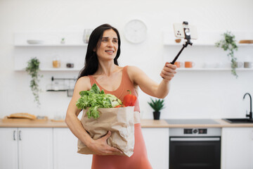 Obraz na płótnie Canvas Cheerful woman in activewear posing at camera of mobile on monopod while holding carrier bag with food. Sports blogger with fresh vegetables and greens for dinner shooting photos after home workout.