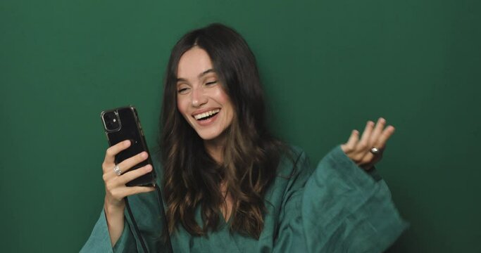 Smiling excited young woman in green dress posing isolated on green background in studio. Pointing on cellphone index finger using mobile phone happy win gesture. Happy curly brunette girl.