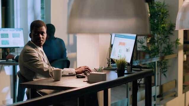 African american expert accountant sitting at desk during nightshift, working on business revenue statistical charts in modern office while colleague in background takes break