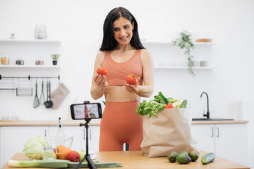 Cheerful brunette sportswoman holding fresh tomatoes while smiling at mobile camera on tripod in...