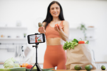 Selective focus of cell phone on tripod being used by cheerful lady with fresh salad ingredients from shopping bag. Fitness and nutrition blogger sharing tips on choosing proper products for meals.
